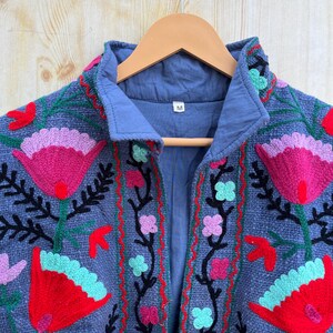 K&ouml;nnte beinhalten: Eine blaue bestickte Jacke mit einem floralen Muster in den Farben Pink, Rot, Gr&uuml;n und Schwarz. Die Jacke hat einen Kragen und einen Knopfverschluss. Das Gr&ouml;&szlig;enetikett zeigt "M" an.