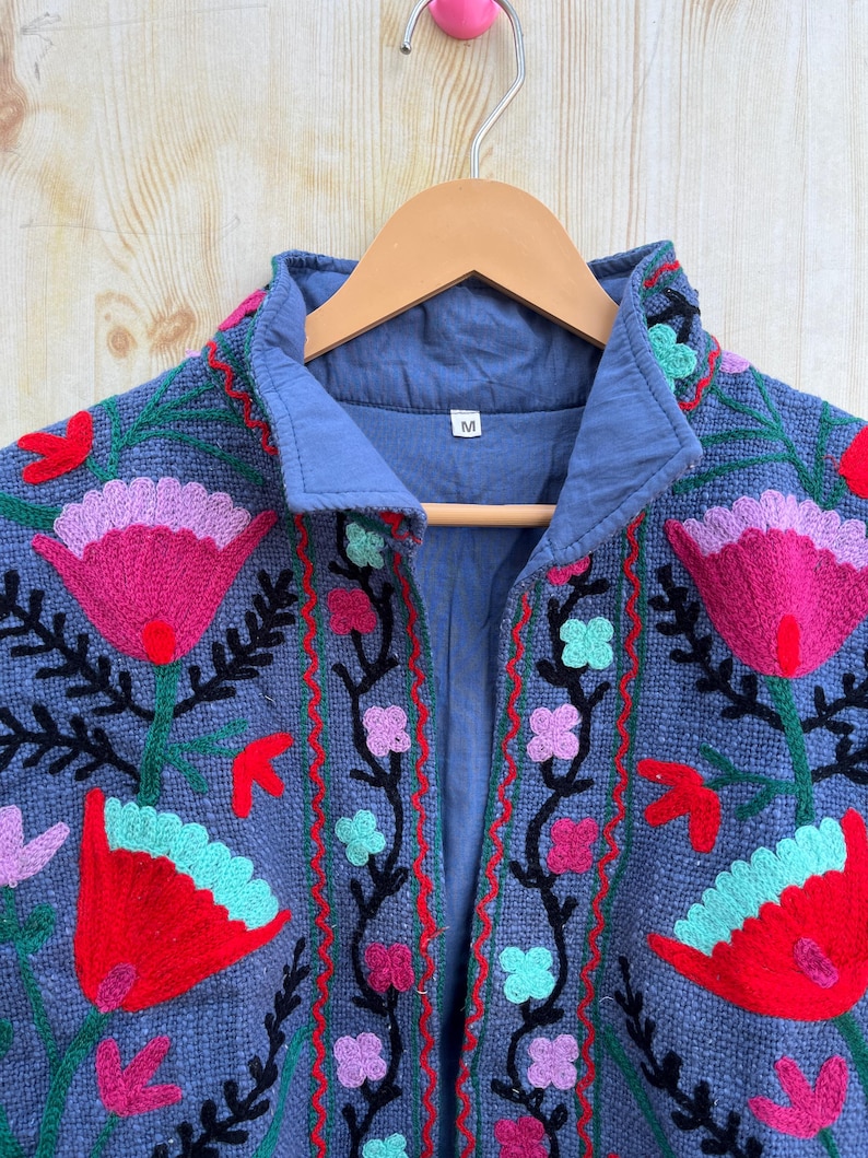K&ouml;nnte beinhalten: Eine blaue bestickte Jacke mit einem floralen Muster in den Farben Pink, Rot, Gr&uuml;n und Schwarz. Die Jacke hat einen Kragen und einen Knopfverschluss. Das Gr&ouml;&szlig;enetikett zeigt "M" an.
