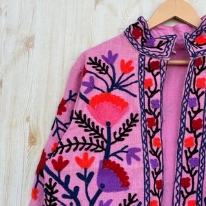 以下が含まれることがあります： ピンク色の刺繍入りジャケット、花柄のデザイン。 ジャケットはスタンドカラーで、赤、紫、黒の刺繍の花と葉で飾られています。 ジャケットは木製のハンガーに掛けられています。