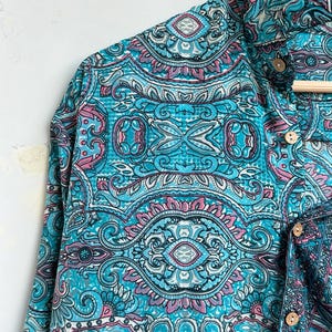 Op de afbeelding: Een shirt met lange mouwen met een levendig paisley-patroon in tinten turquoise, roze en wit. Het heeft een button-down kraag en houten knopen. De stof ziet er zacht en vloeiend uit.