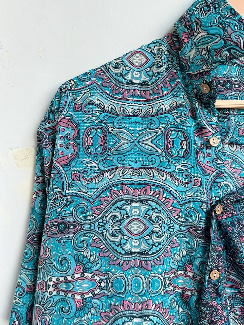 Op de afbeelding: Een shirt met lange mouwen met een levendig paisley-patroon in tinten turquoise, roze en wit. Het heeft een button-down kraag en houten knopen. De stof ziet er zacht en vloeiend uit.