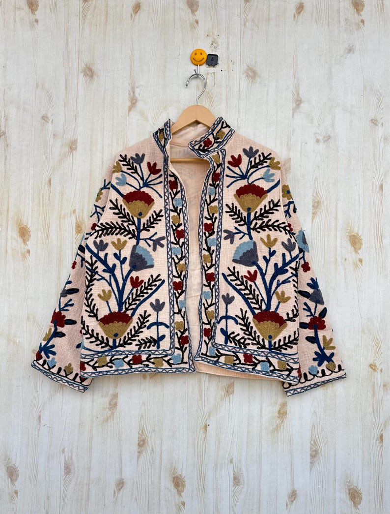 Pode incluir: Casaco bordado rosa claro com um design floral em tons de azul, vermelho e dourado. O casaco tem uma gola alta e est&aacute; pendurado em um cabide de madeira contra um fundo de madeira clara.