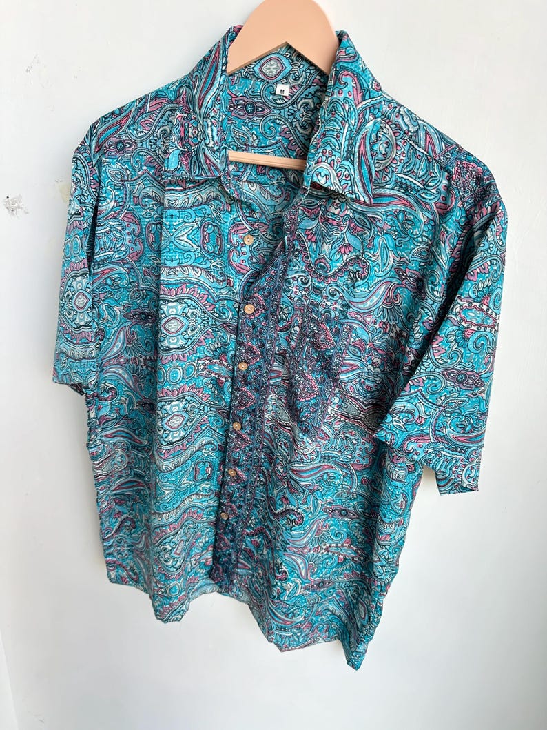 Op de afbeelding: Een overhemd met korte mouwen en een levendig paisley-patroon in tinten turquoise, roze en blauw. Het overhemd hangt aan een houten kleerhanger. De stof lijkt lichtgewicht en geschikt voor warm weer.