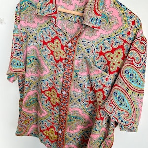 Op de afbeelding: Een shirt met korte mouwen met een levendig paisley patroon in roze, rood, blauw en geel. Het shirt hangt aan een houten hanger en toont het ingewikkelde ontwerp en kleurenpalet. Het heeft een klassieke kraag en een knoopsluiting.