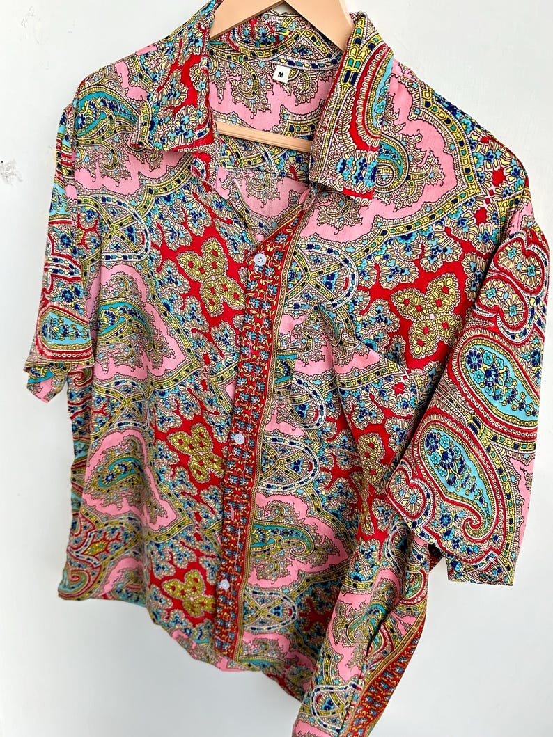 Op de afbeelding: Een shirt met korte mouwen met een levendig paisley patroon in roze, rood, blauw en geel. Het shirt hangt aan een houten hanger en toont het ingewikkelde ontwerp en kleurenpalet. Het heeft een klassieke kraag en een knoopsluiting.