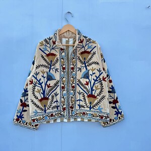 Pu&ograve; includere: Una giacca color crema con un ricamo floreale in tonalit&agrave; di blu, rosso e marrone. La giacca ha una chiusura a zip e maniche lunghe.