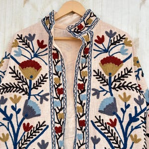Pode incluir: Casaco rosa claro bordado com gola mandarim, pendurado num cabide de madeira. O casaco apresenta um desenho floral em fio azul, vermelho, amarelo e preto. O casaco &eacute; aberto na frente.