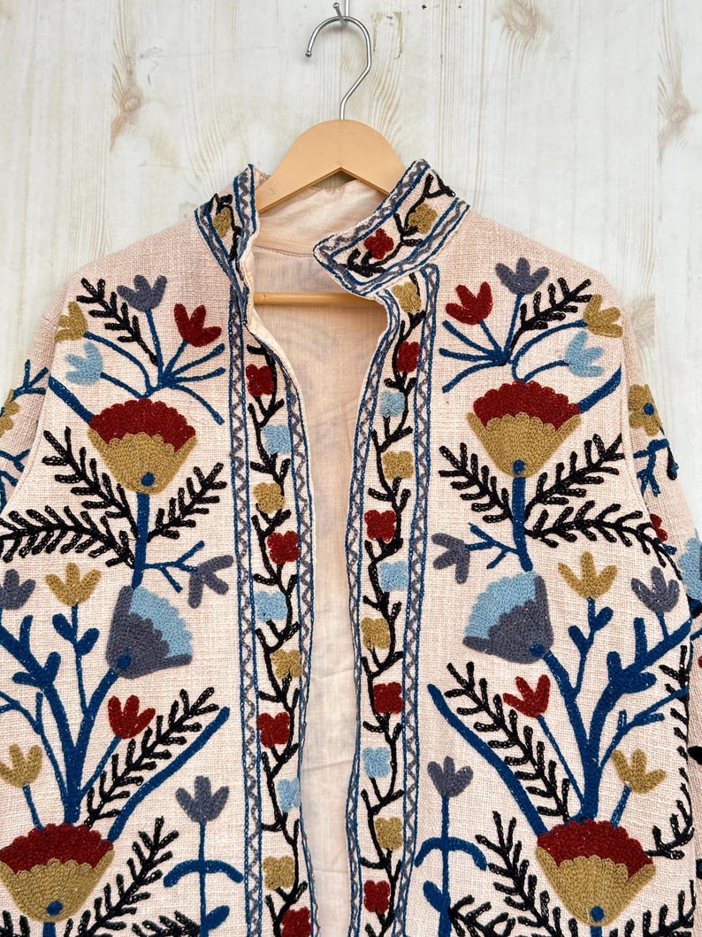 Pode incluir: Casaco rosa claro bordado com gola mandarim, pendurado num cabide de madeira. O casaco apresenta um desenho floral em fio azul, vermelho, amarelo e preto. O casaco &eacute; aberto na frente.