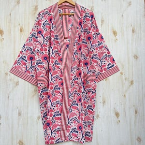 K&ouml;nnte beinhalten: Kimono-Robe mit floralem Muster in Rosa, Wei&szlig; und Marineblau. Das Muster wiederholt Blumen und Bl&auml;tter. Die Robe h&auml;ngt an einem Holzb&uuml;gel vor einem hellen Holzgrund.