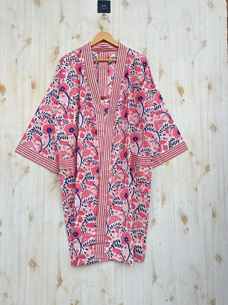 K&ouml;nnte beinhalten: Kimono-Robe mit floralem Muster in Rosa, Wei&szlig; und Marineblau. Das Muster wiederholt Blumen und Bl&auml;tter. Die Robe h&auml;ngt an einem Holzb&uuml;gel vor einem hellen Holzgrund.