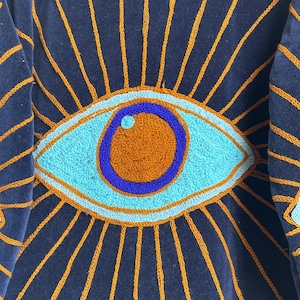 May include: Close-up of a navy blue textile with an embroidered eye design. The eye is turquoise, orange, and blue, surrounded by orange sun rays. The textile has a soft, velvety texture.