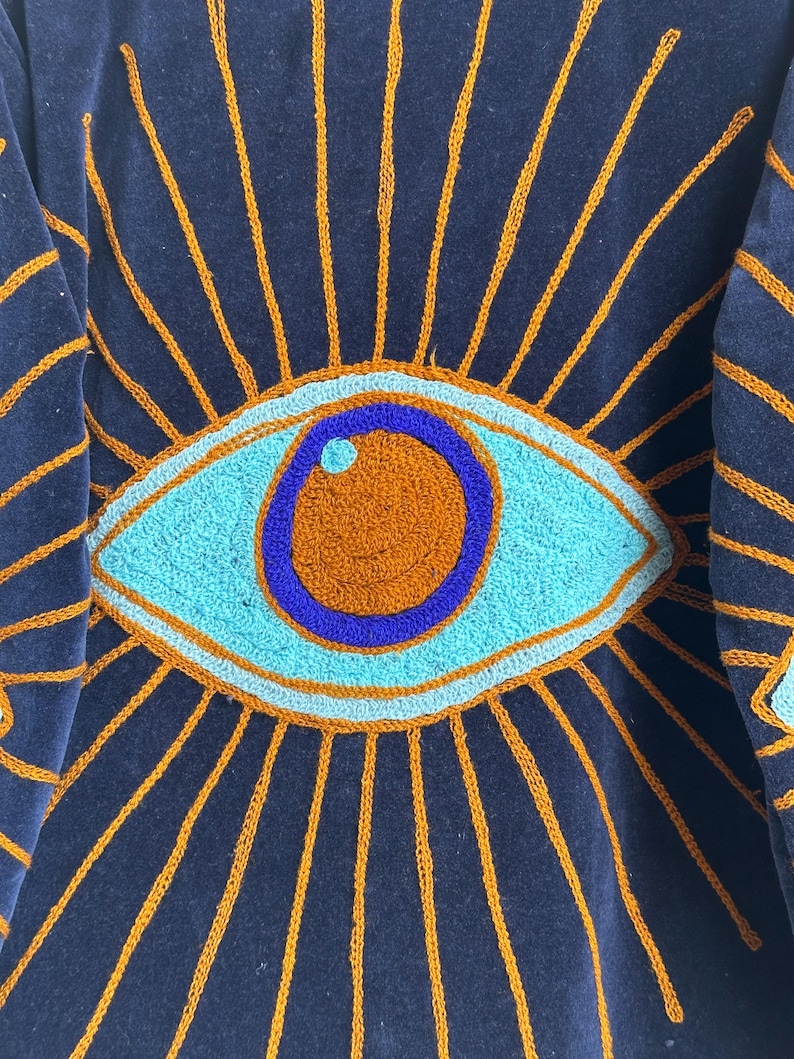 May include: Close-up of a navy blue textile with an embroidered eye design. The eye is turquoise, orange, and blue, surrounded by orange sun rays. The textile has a soft, velvety texture.
