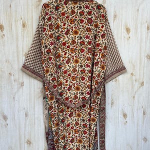 May include: A floral print kimono robe with a cream background and red, yellow, and green flowers. The robe has long sleeves with a geometric pattern on the cuffs and a matching belt. The robe is hanging on a wooden hanger against a light wood background.