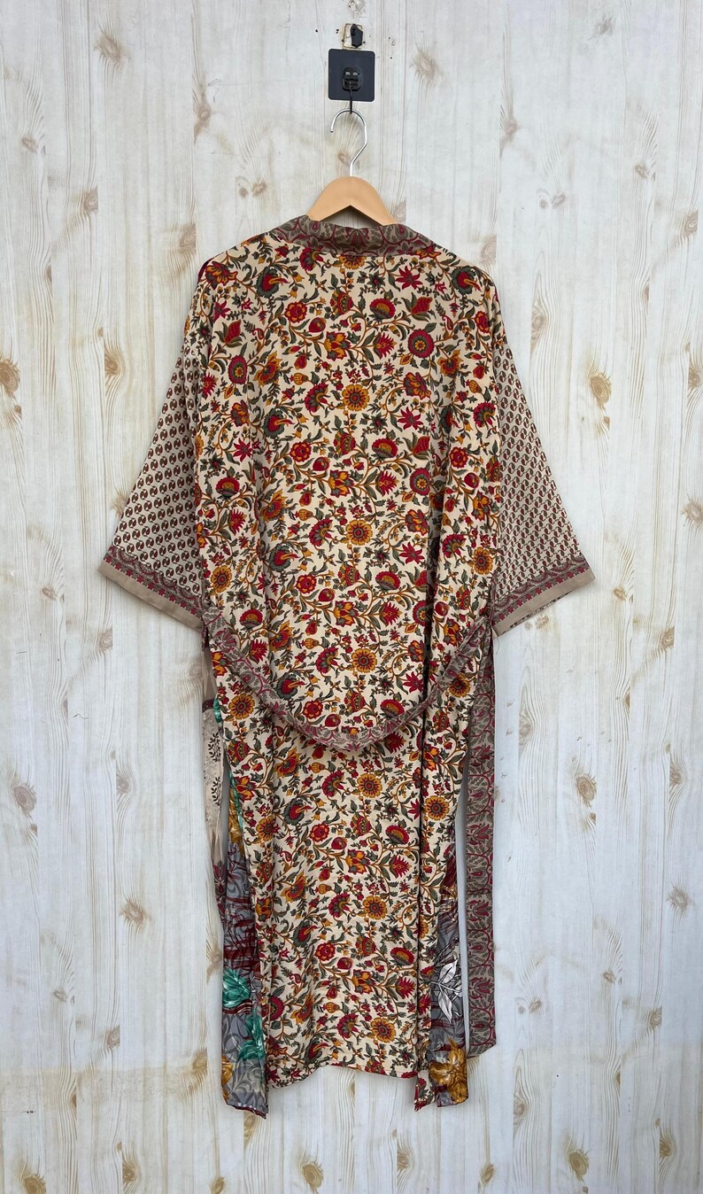 May include: A floral print kimono robe with a cream background and red, yellow, and green flowers. The robe has long sleeves with a geometric pattern on the cuffs and a matching belt. The robe is hanging on a wooden hanger against a light wood background.
