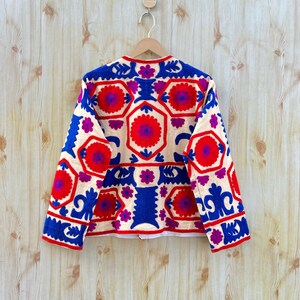May include: A cream-coloured jacket with a vibrant floral pattern in red, blue, and purple. The jacket features a round neckline and long sleeves. The floral design is embroidered across the jacket's front and back.