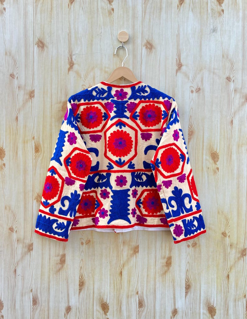 May include: A cream-coloured jacket with a vibrant floral pattern in red, blue, and purple. The jacket features a round neckline and long sleeves. The floral design is embroidered across the jacket's front and back.