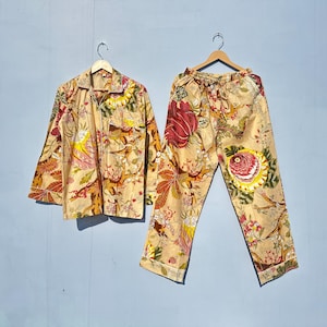 Op de afbeelding: Een beige pyjamaset met een bloemenprint met rode, gele en groene bloemen. Het bovenstuk heeft een knoopsluiting aan de voorkant en lange mouwen. De broek heeft een elastische tailleband.