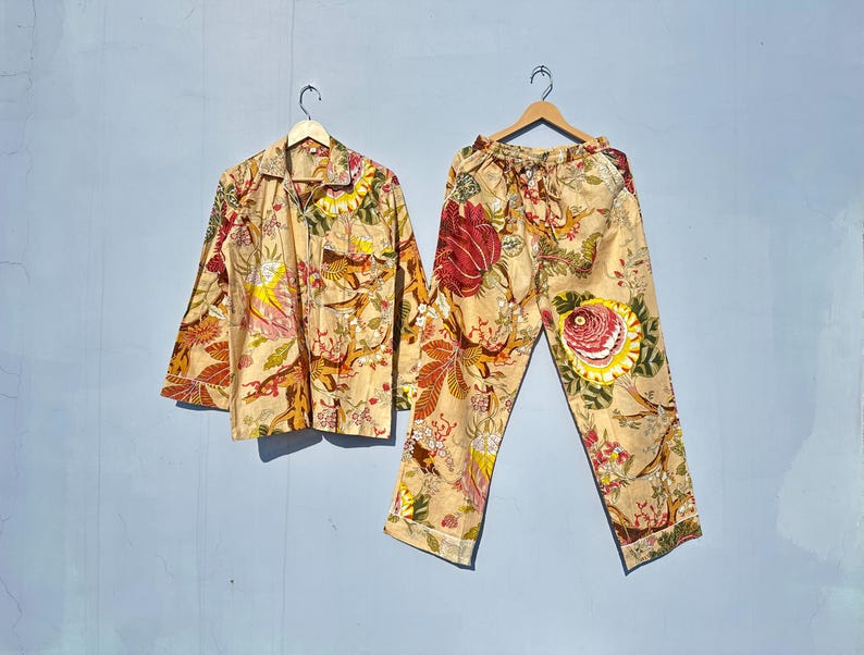 Op de afbeelding: Een beige pyjamaset met een bloemenprint met rode, gele en groene bloemen. Het bovenstuk heeft een knoopsluiting aan de voorkant en lange mouwen. De broek heeft een elastische tailleband.