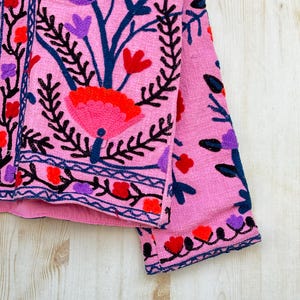 以下が含まれることがあります： ピンク色の刺繍入りジャケット、花柄のデザイン。 赤、紫、ピンクの花と黒と青のアクセントが特徴です。 刺繍は細かく、ジャケットの前面と袖を覆っています。