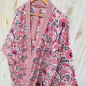 K&ouml;nnte beinhalten: Ein Bademantel mit floralem Muster in Rosa, Marineblau und Wei&szlig; h&auml;ngt an einem Holzb&uuml;gel. Der Bademantel zeigt ein sich wiederholendes Muster aus gro&szlig;en rosa Blumen und marineblauen Bl&auml;ttern auf wei&szlig;em Grund. Die Kanten sind mit rosa und wei&szlig;en Streifen verziert.