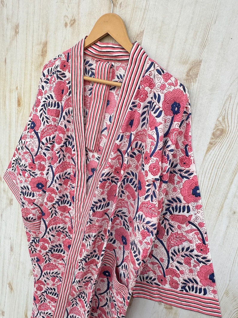 K&ouml;nnte beinhalten: Ein Bademantel mit floralem Muster in Rosa, Marineblau und Wei&szlig; h&auml;ngt an einem Holzb&uuml;gel. Der Bademantel zeigt ein sich wiederholendes Muster aus gro&szlig;en rosa Blumen und marineblauen Bl&auml;ttern auf wei&szlig;em Grund. Die Kanten sind mit rosa und wei&szlig;en Streifen verziert.