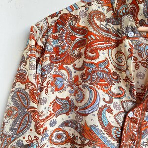 Op de afbeelding: Cr&egrave;mekleurig overhemd met knopen en een levendig paisley patroon in tinten oranje, blauw en bruin. Het overhemd heeft een klassieke kraag en manchetten met knopen. De stof heeft een zijdeachtige glans.