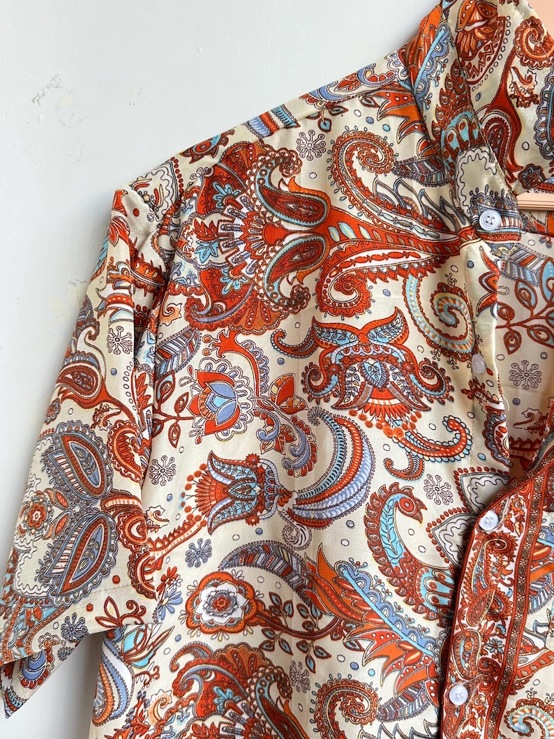 Op de afbeelding: Cr&egrave;mekleurig overhemd met knopen en een levendig paisley patroon in tinten oranje, blauw en bruin. Het overhemd heeft een klassieke kraag en manchetten met knopen. De stof heeft een zijdeachtige glans.