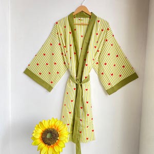 May include: A light green and white striped robe with red heart accents, hanging on a wooden hanger. The robe has olive green trim and a matching belt. A yellow sunflower is in the foreground.