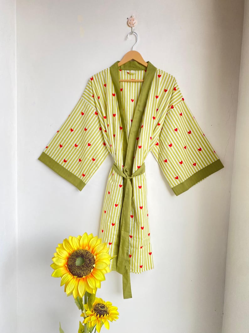 May include: A light green and white striped robe with red heart accents, hanging on a wooden hanger. The robe has olive green trim and a matching belt. A yellow sunflower is in the foreground.