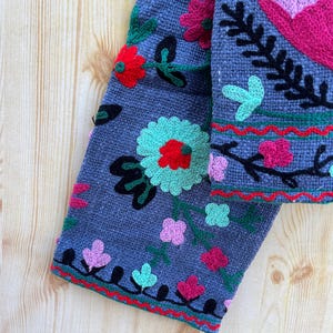 K&ouml;nnte beinhalten: Ein grauer Stoff&auml;rmel mit bunten gestickten Blumen und Bl&auml;ttern. Der &Auml;rmel hat einen roten, gr&uuml;nen und schwarzen Besatz.