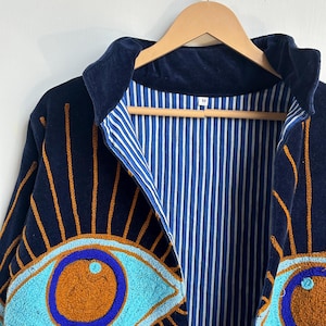 May include: Navy blue jacket with embroidered eye designs in turquoise and orange. The jacket has a blue and white striped lining and a dark blue velvet collar. The jacket is hanging on a wooden hanger.
