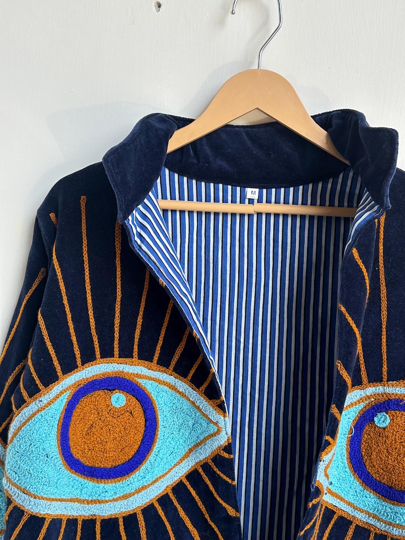 May include: Navy blue jacket with embroidered eye designs in turquoise and orange. The jacket has a blue and white striped lining and a dark blue velvet collar. The jacket is hanging on a wooden hanger.