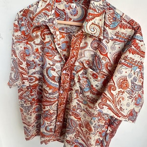 Op de afbeelding: Een cr&egrave;mekleurig overhemd met een paisley patroon in rood, oranje en blauw. Het overhemd hangt aan een houten hanger.