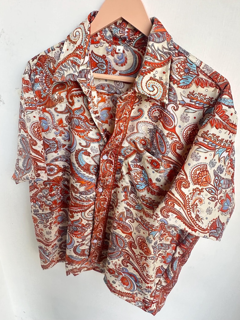 Op de afbeelding: Een cr&egrave;mekleurig overhemd met een paisley patroon in rood, oranje en blauw. Het overhemd hangt aan een houten hanger.