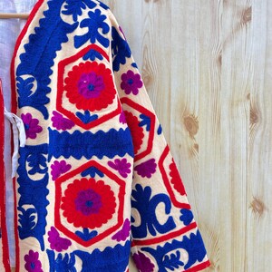 May include: Embroidered cream jacket with a floral design in red, blue, and purple. The jacket showcases detailed stitching along the sleeves and edges. The garment's design incorporates geometric and floral motifs, creating a visually rich texture.