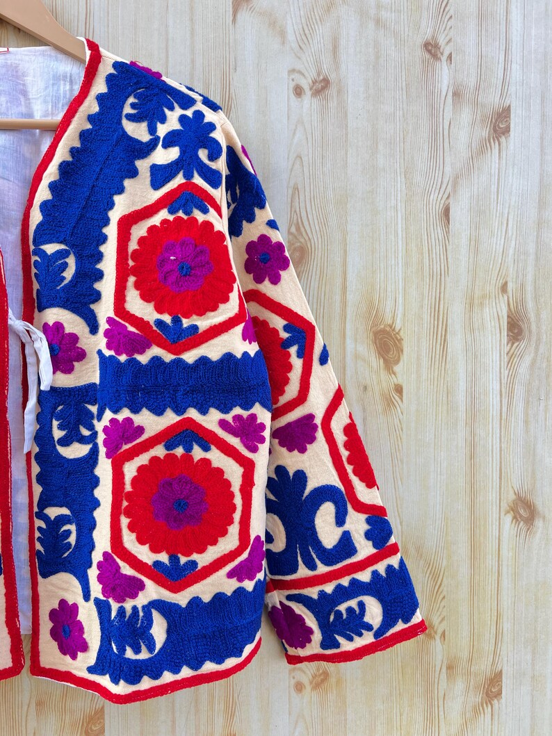 May include: Embroidered cream jacket with a floral design in red, blue, and purple. The jacket showcases detailed stitching along the sleeves and edges. The garment's design incorporates geometric and floral motifs, creating a visually rich texture.