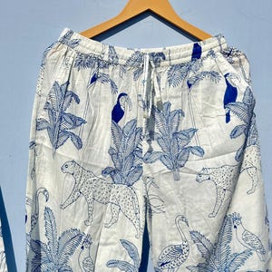 May include: White cotton trousers with a blue tropical print featuring toucans, leopards, and flamingos. The trousers have an elastic waistband and a drawstring.