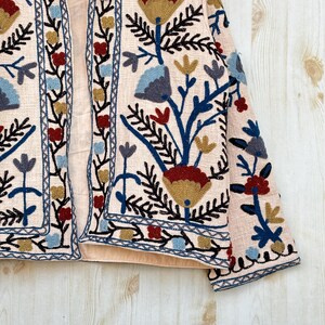 Pode incluir: Casaco rosa claro bordado com um design floral. O casaco apresenta uma base creme com bordados intrincados em vermelho, azul, dourado e preto. O padr&atilde;o floral inclui flores e folhas estilizadas, criando um visual detalhado e art&iacute;stico.