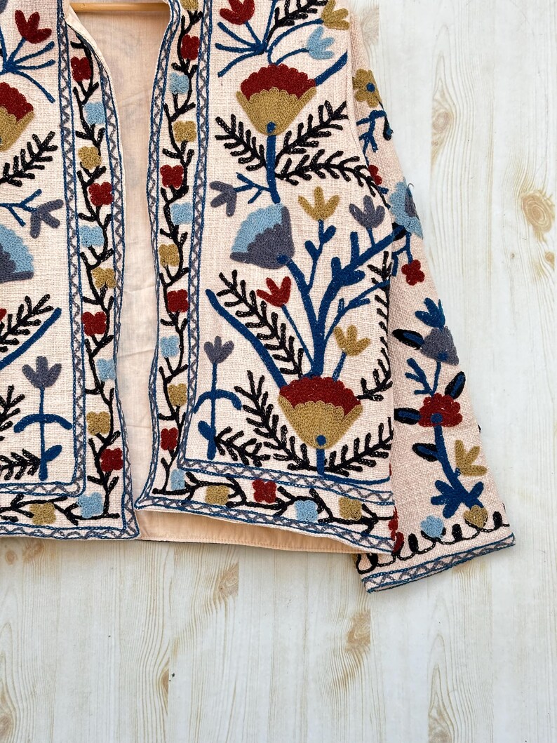 Pode incluir: Casaco rosa claro bordado com um design floral. O casaco apresenta uma base creme com bordados intrincados em vermelho, azul, dourado e preto. O padr&atilde;o floral inclui flores e folhas estilizadas, criando um visual detalhado e art&iacute;stico.