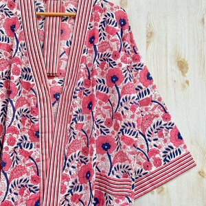 K&ouml;nnte beinhalten: Ein Bademantel mit floralem Muster in Rosa, Wei&szlig; und Marineblau, der an einem Holzb&uuml;gel h&auml;ngt. Der Bademantel hat ein sich wiederholendes Muster aus Blumen und Bl&auml;ttern. Die Verzierungen und Kanten sind mit Streifen versehen.