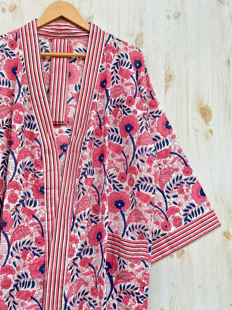 K&ouml;nnte beinhalten: Ein Bademantel mit floralem Muster in Rosa, Wei&szlig; und Marineblau, der an einem Holzb&uuml;gel h&auml;ngt. Der Bademantel hat ein sich wiederholendes Muster aus Blumen und Bl&auml;ttern. Die Verzierungen und Kanten sind mit Streifen versehen.