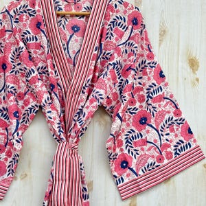 K&ouml;nnte beinhalten: Ein Bademantel mit floralem Muster in Rosa, Wei&szlig; und Marineblau h&auml;ngt an einem Holzb&uuml;gel. Der Bademantel hat ein sich wiederholendes Muster aus Blumen und Bl&auml;ttern. Der Bademantel hat eine gestreifte Bord&uuml;re und einen passenden G&uuml;rtel.