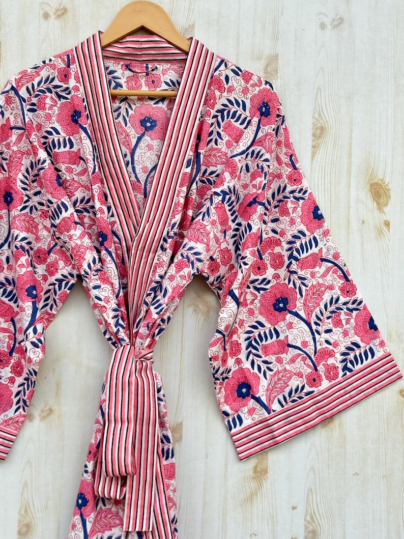 K&ouml;nnte beinhalten: Ein Bademantel mit floralem Muster in Rosa, Wei&szlig; und Marineblau h&auml;ngt an einem Holzb&uuml;gel. Der Bademantel hat ein sich wiederholendes Muster aus Blumen und Bl&auml;ttern. Der Bademantel hat eine gestreifte Bord&uuml;re und einen passenden G&uuml;rtel.