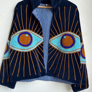 May include: Navy blue velvet jacket with embroidered eye designs in turquoise, brown, and gold. The jacket features a high collar, a striped blue and white lining, and a zip-up front. The eye designs are on the front and sleeves.