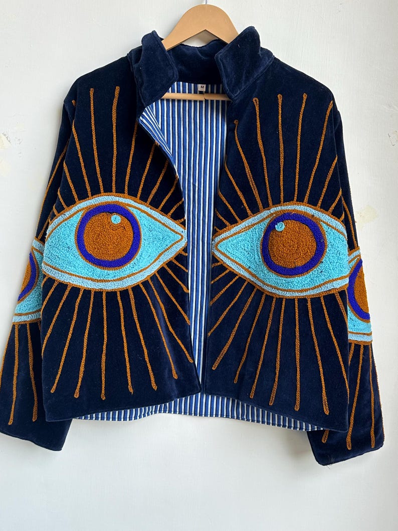 May include: Navy blue velvet jacket with embroidered eye designs in turquoise, brown, and gold. The jacket features a high collar, a striped blue and white lining, and a zip-up front. The eye designs are on the front and sleeves.