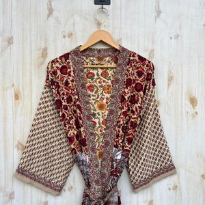 May include: A floral print robe with a beige background and red, yellow, and green floral patterns. The robe has wide sleeves and a matching belt. The robe is hanging on a wooden hanger.