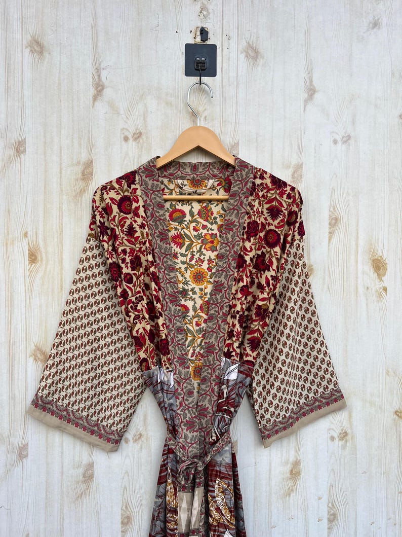 May include: A floral print robe with a beige background and red, yellow, and green floral patterns. The robe has wide sleeves and a matching belt. The robe is hanging on a wooden hanger.