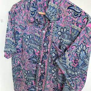 Op de afbeelding: Een overhemd met knopen en korte mouwen met een levendig paisley-patroon in roze, blauw en grijs. Het overhemd hangt aan een houten kleerhanger. De stof heeft een gedetailleerd bloemen- en abstract ontwerp.