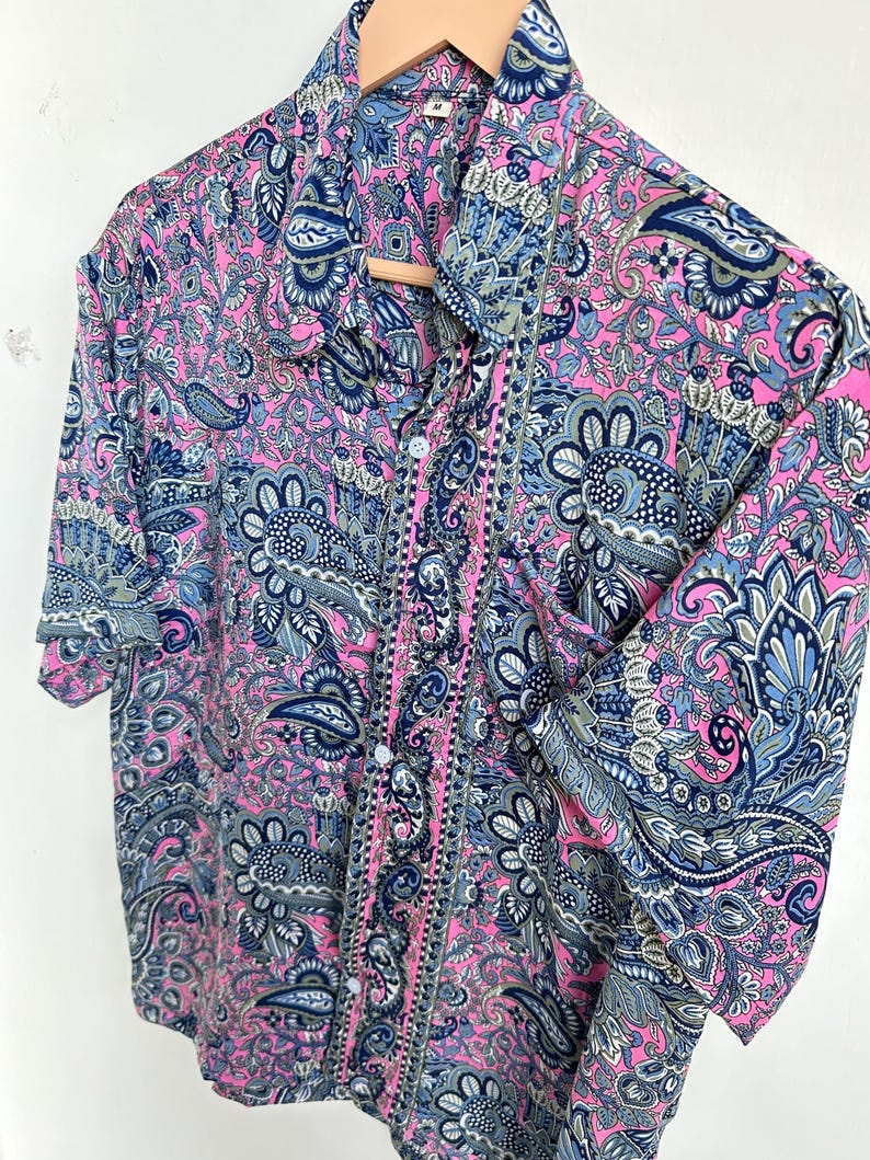 Op de afbeelding: Een overhemd met knopen en korte mouwen met een levendig paisley-patroon in roze, blauw en grijs. Het overhemd hangt aan een houten kleerhanger. De stof heeft een gedetailleerd bloemen- en abstract ontwerp.