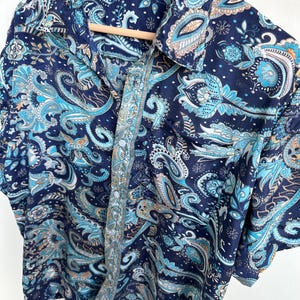 Op de afbeelding: Een overhemd met korte mouwen met een paisley patroon in marineblauw, lichtblauw en bruin. Het overhemd hangt aan een houten hanger. Het heeft een kraag en een knoopsluiting. Maat M.